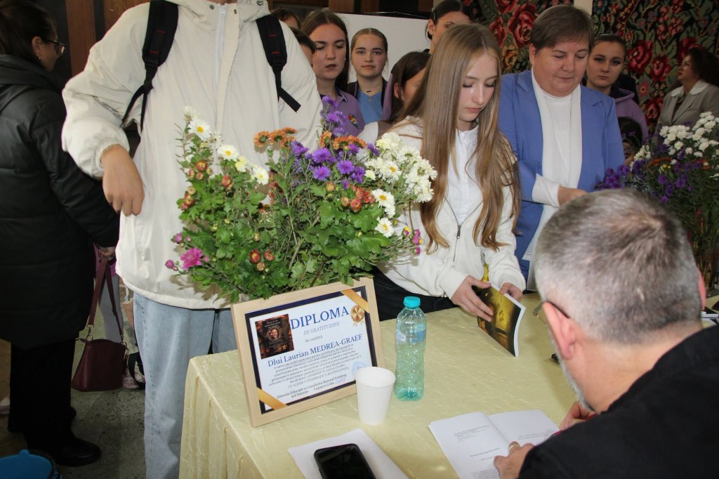 Un exemplar al volumului este semnat la masa cu flori și diploma de gratitudine, în timp ce copiii și ceilalți participanți așteaptă.
