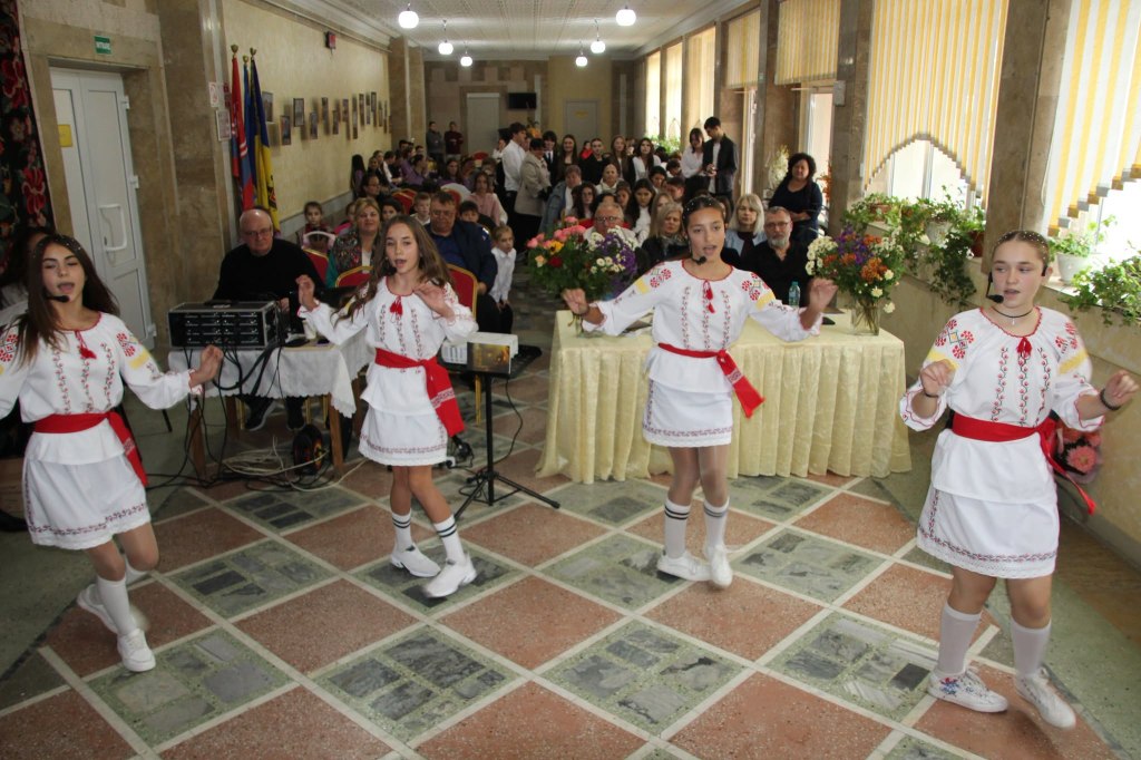 Patru fete în costume albe cu motive roșii dansează în fața publicului din sala de la Slobozia-Dușca.