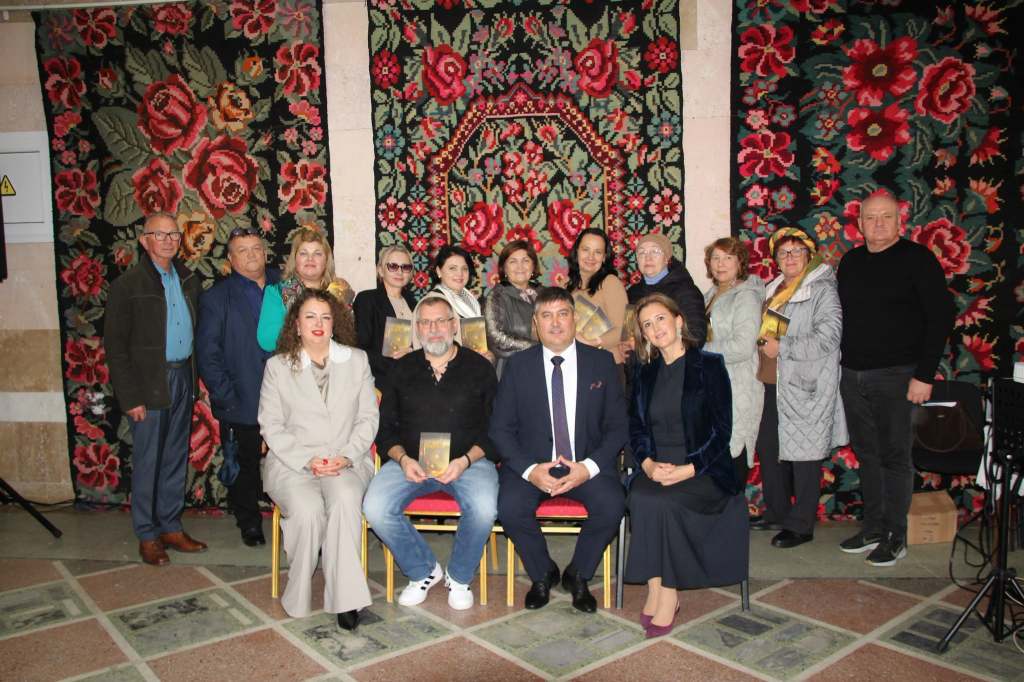 Participanții la lansarea volumului „Ephemer” pozează împreună în fața covoarelor tradiționale cu motive florale.