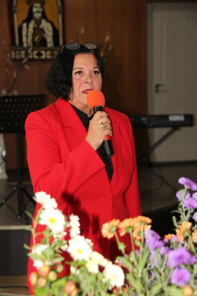 O participantă în ținută roșie vorbește la microfon, lângă aranjamentele florale din sala evenimentului.